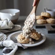 healthy biscuits and gravy on a white plate with a fork on the plate and a woman putting more gravy on the biscuits with a wooden spoon, all on a gray table