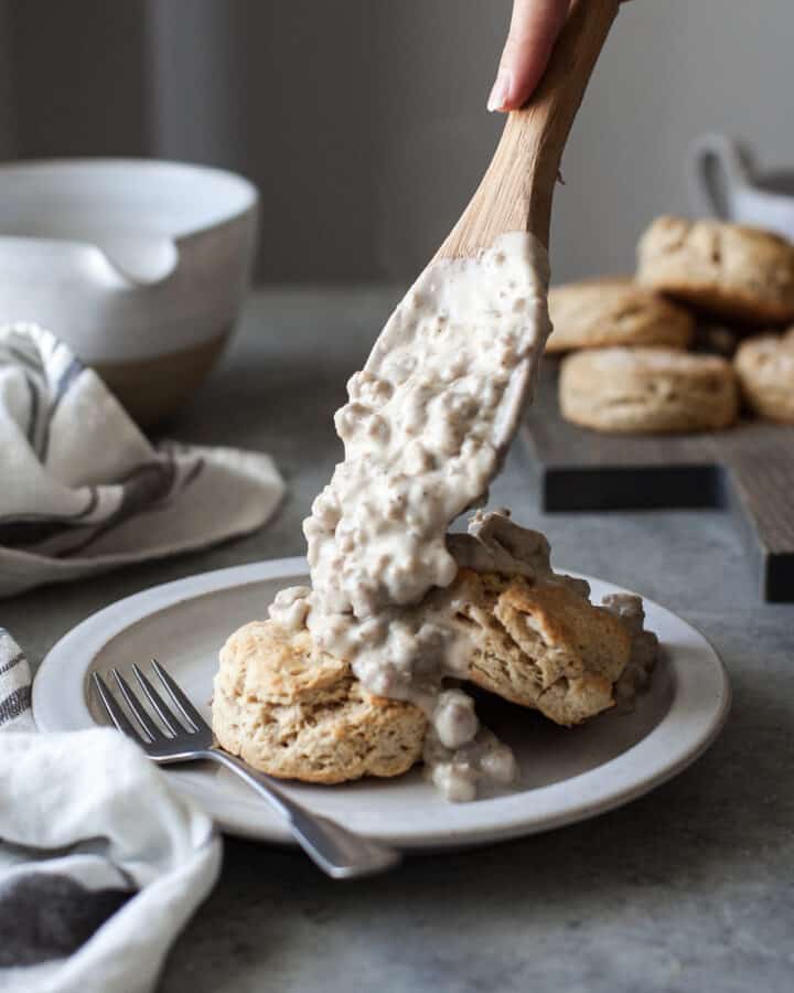 healthy biscuits and gravy on a white plate with a fork on the plate and a woman putting more gravy on the biscuits with a wooden spoon, all on a gray table