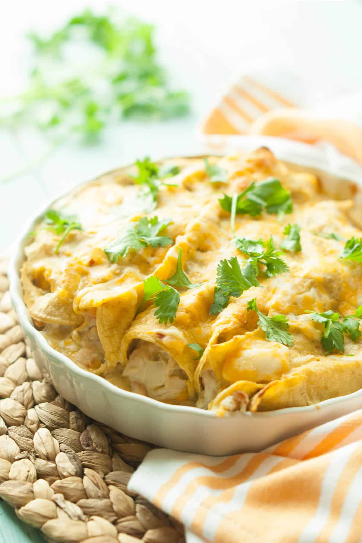 Cream cheese gluten-free chicken enchiladas in a white pan with cilantro on top on a rattan mat with a white and orange linen napkin.