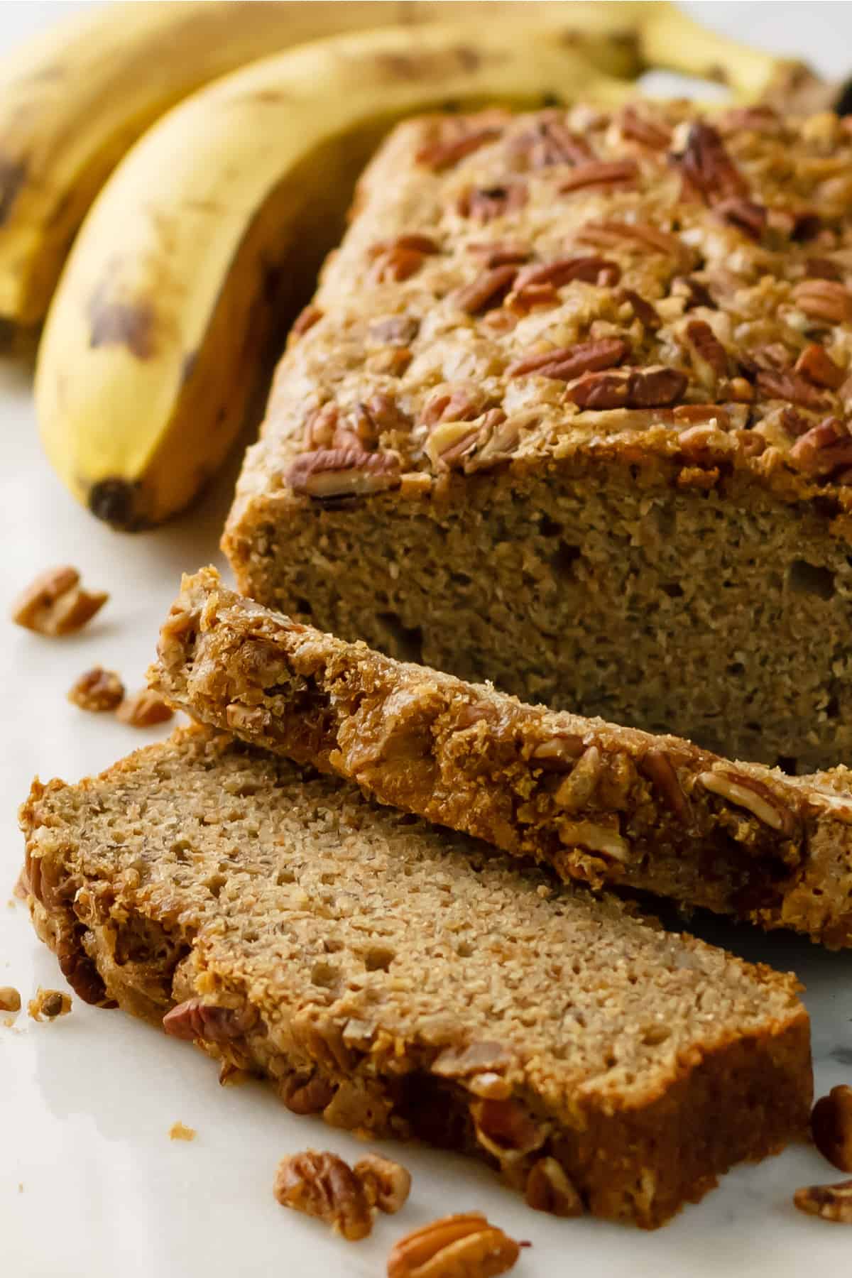 Dairy-free banana bread recipe sliced with ripe bananas in the background on a white table.