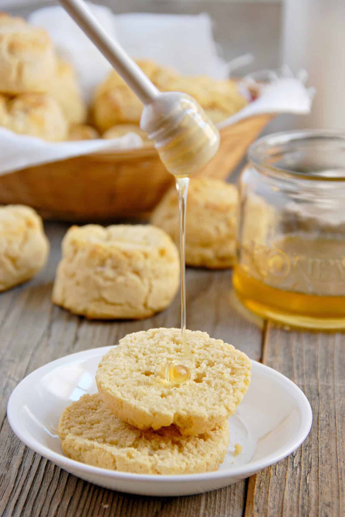 Fluffy Paleo biscuits on a small white plate with honey being drizzled on them and some behind in a basket and a small jar of honey in the background.