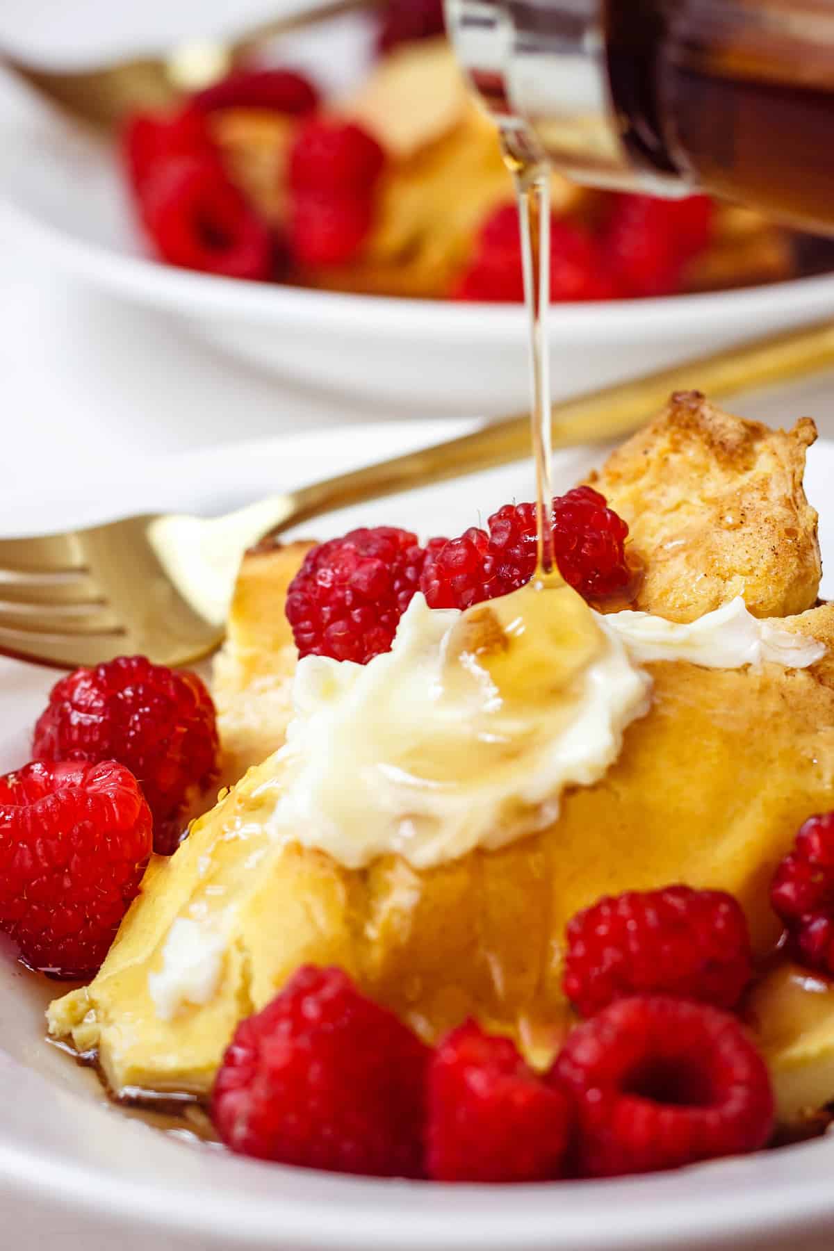 Gluten-free German pancakes on a white plate with raspberries and butter on top and maple syrup being drizzled on top.