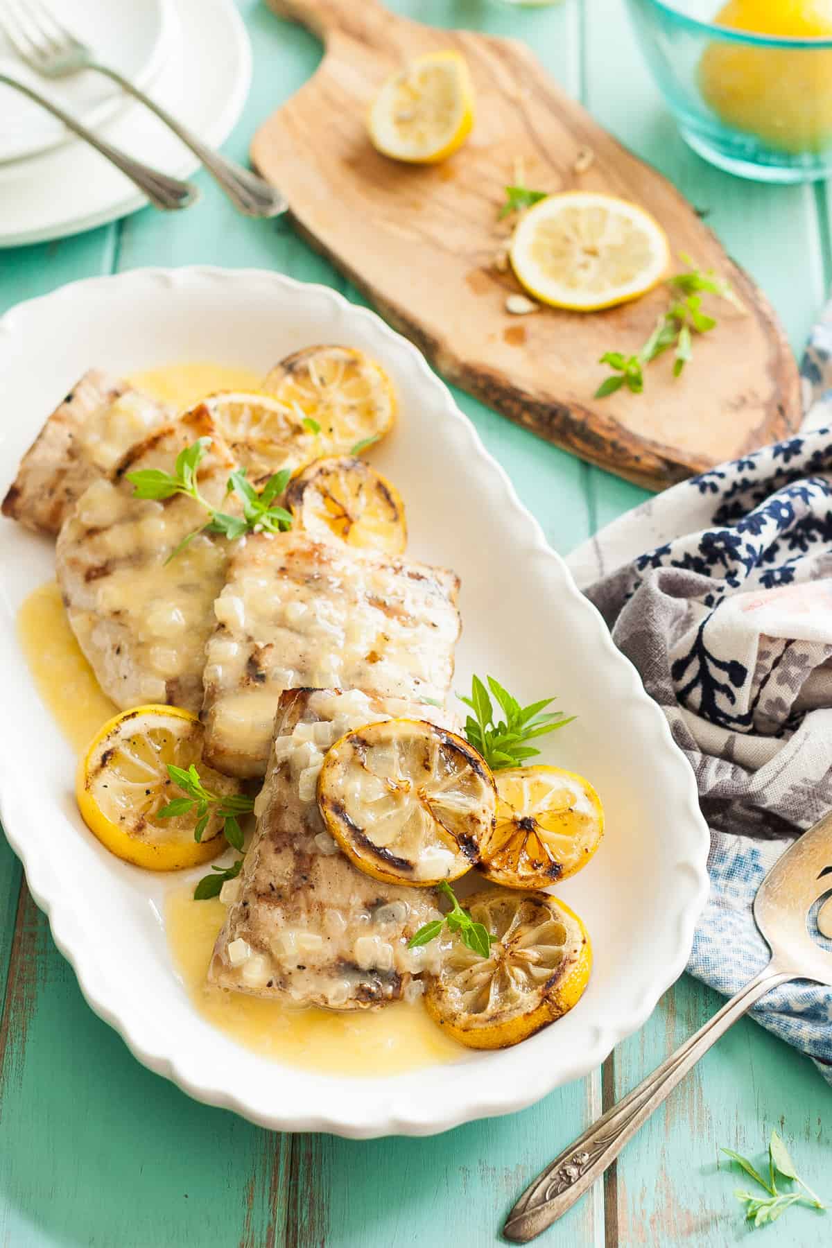 Grilled Mahi Mahi recipe with a lemon butter sauce with sliced lemons on top on a white plate with a turquoise background and a white and blue napkin on the side.
