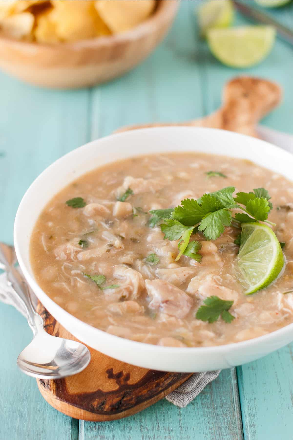 Healthy white chicken chili in a white bowl on a green background on a brown cutting board with a spoon, topped with lime and cilantro.